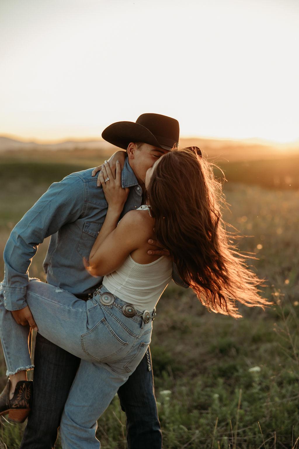 A couple sits in a grassy field at sunset. The woman, laughing and wearing a black hat,. They both wear casual, light-colored clothing during their western engagement photos