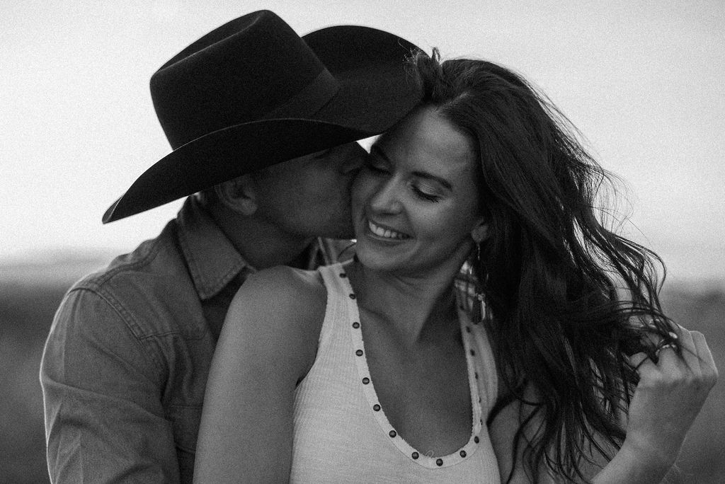 A couple sits in a grassy field at sunset. The woman, laughing and wearing a black hat,. They both wear casual, light-colored clothing during their western engagement photos