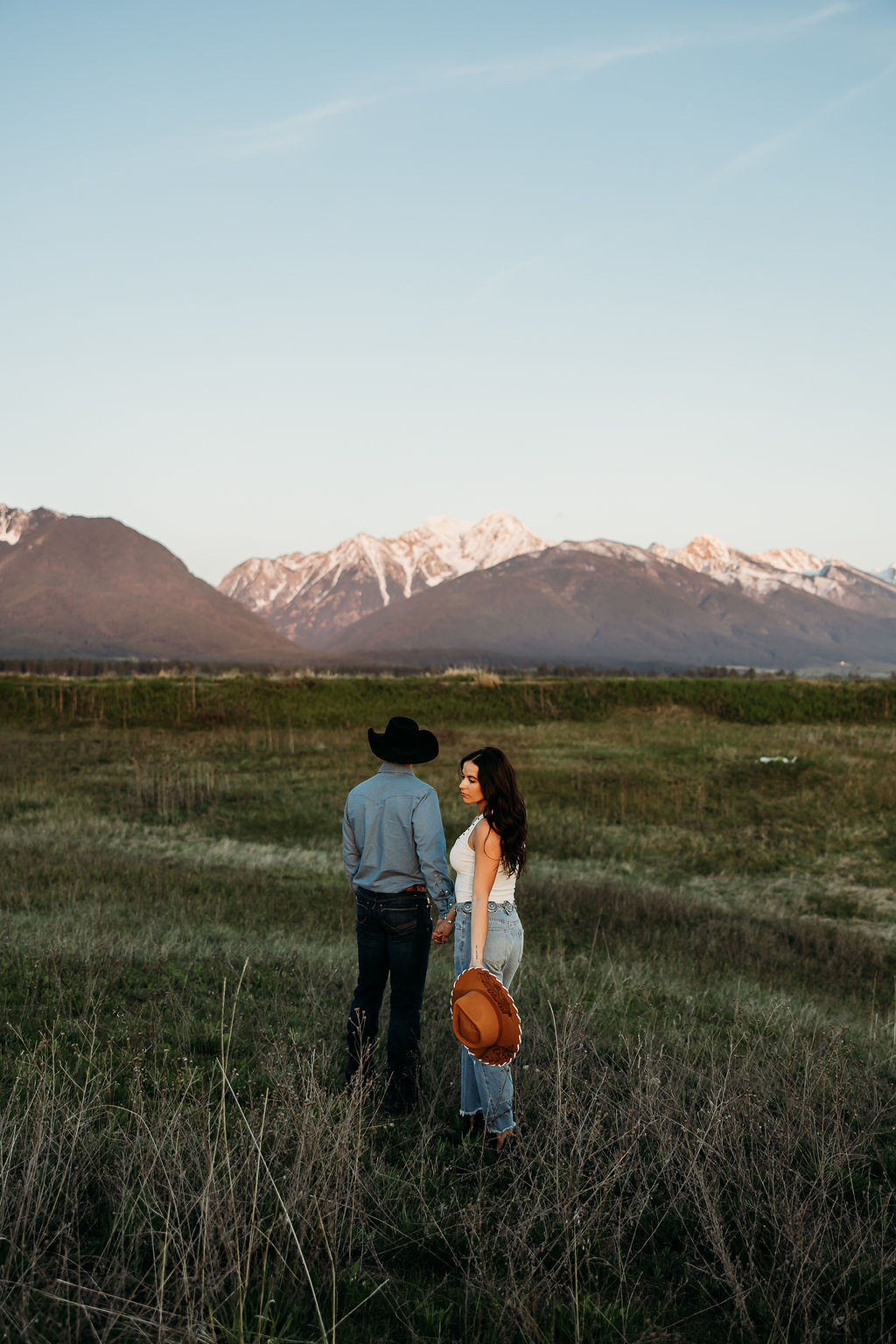 A couple sits in a grassy field at sunset. The woman, laughing and wearing a black hat,. They both wear casual, light-colored clothing during their western engagement photos