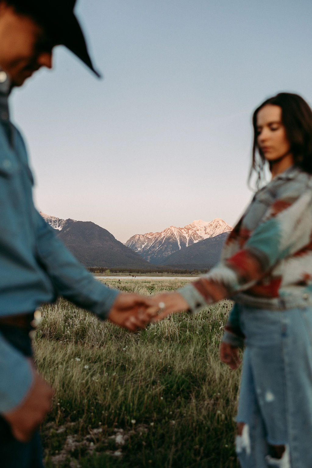 A couple sits in a grassy field at sunset. The woman, laughing and wearing a black hat,. They both wear casual, light-colored clothing during their western engagement photos