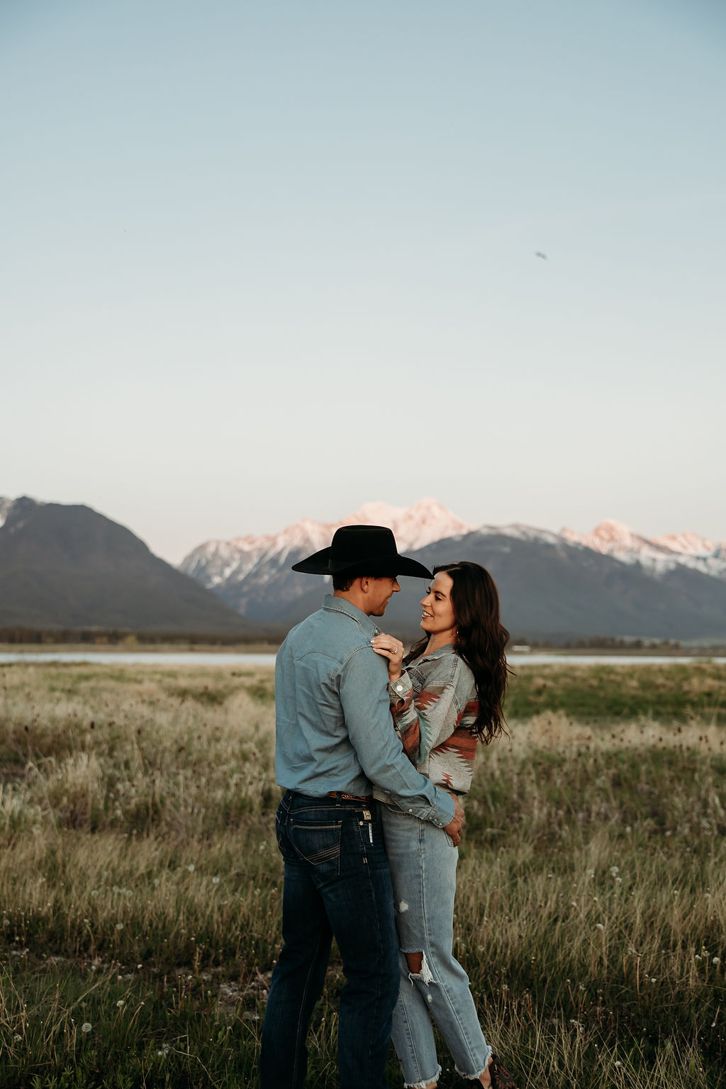 A couple sits in a grassy field at sunset. The woman, laughing and wearing a black hat,. They both wear casual, light-colored clothing during their western engagement photos