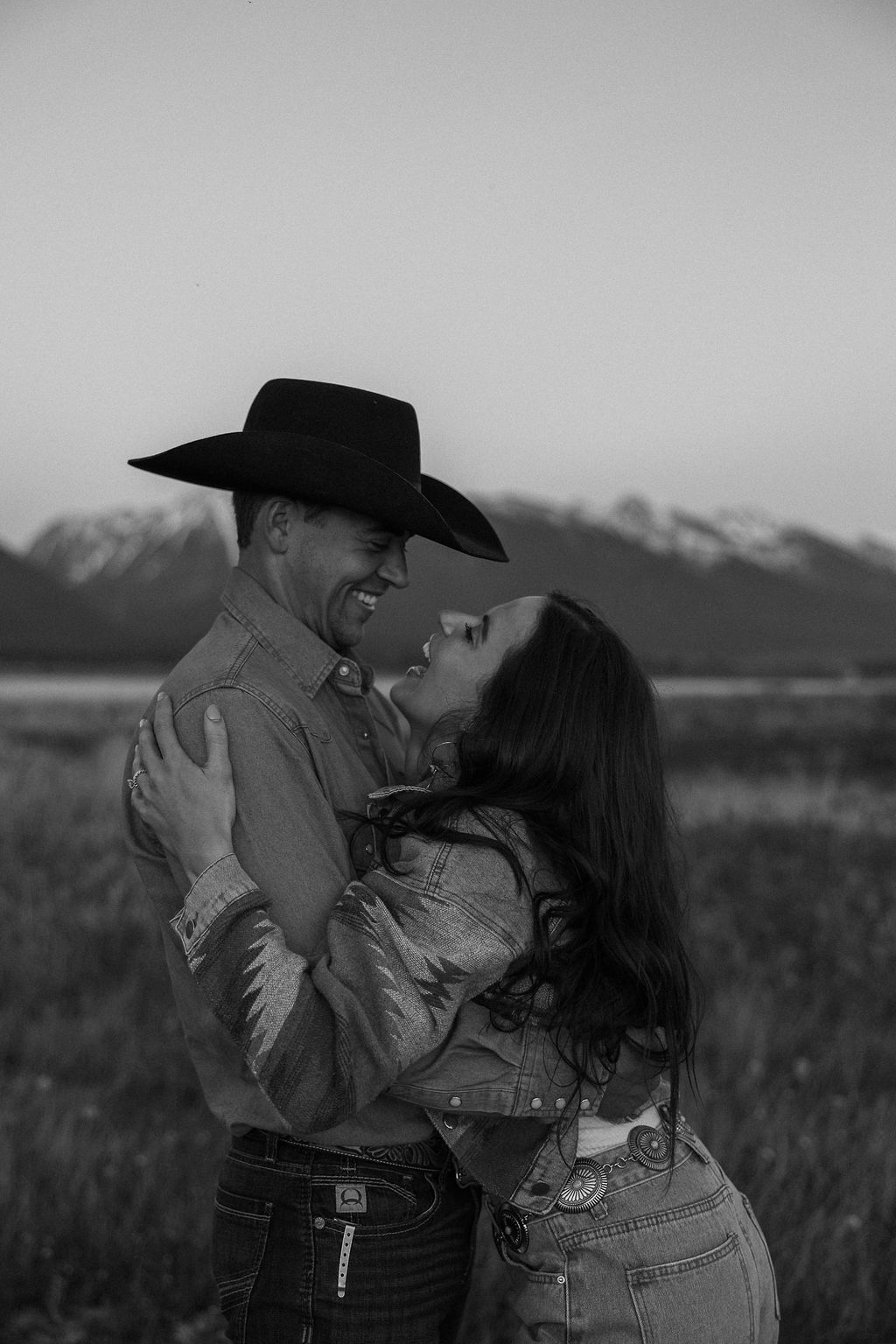 A couple sits in a grassy field at sunset. The woman, laughing and wearing a black hat,. They both wear casual, light-colored clothing during their western engagement photos