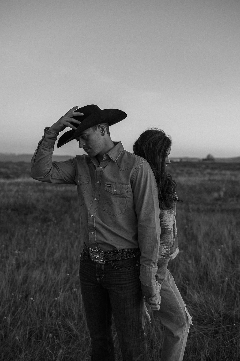 A couple sits in a grassy field at sunset. The woman, laughing and wearing a black hat,. They both wear casual, light-colored clothing during their western engagement photos