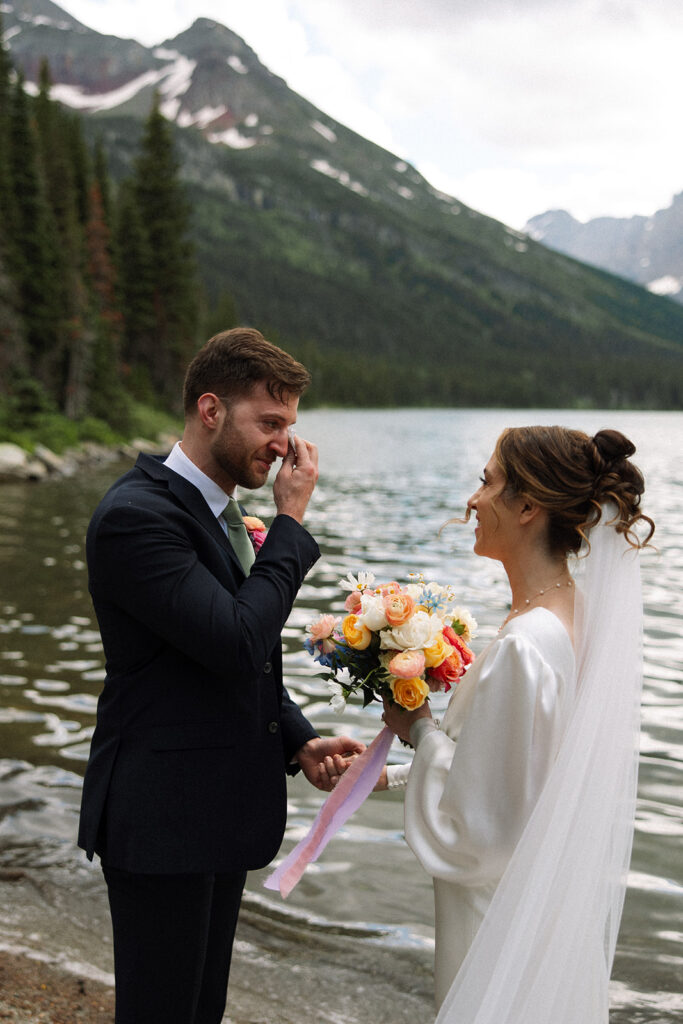 Bride and groom standing by Josephine Lake shoreline surrounded by mountains.
