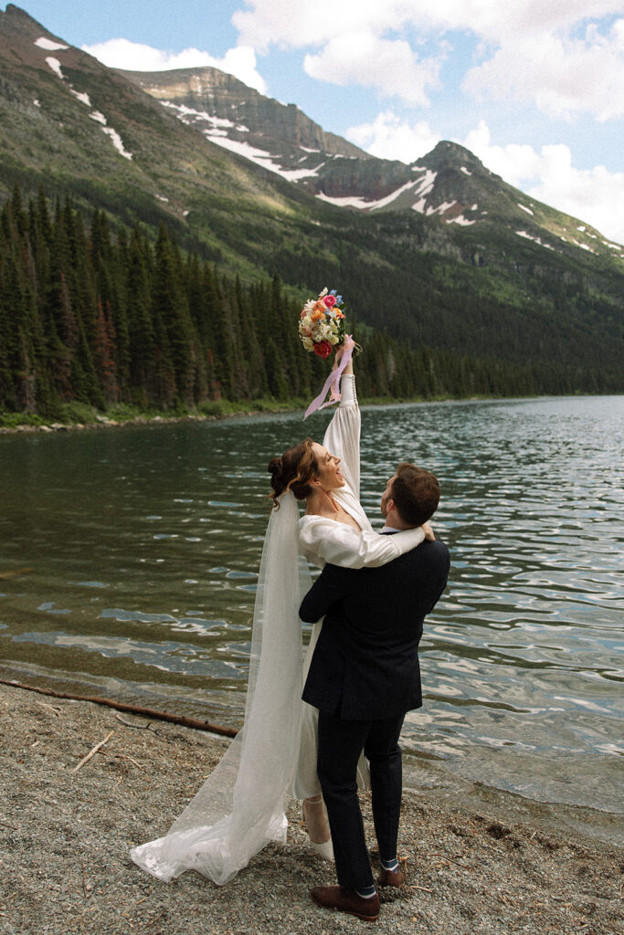 Bride and groom standing by Josephine Lake shoreline surrounded by mountains.
