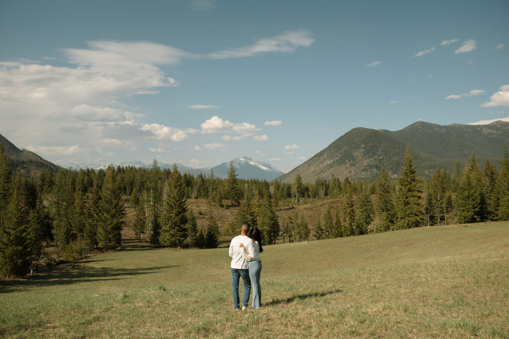 Couple walking through lush green valley with snow-capped mountains during a Glacier National Park engagement session

