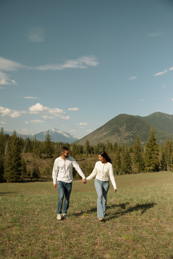 Couple walking through lush green valley with snow-capped mountains during a Glacier National Park engagement session
