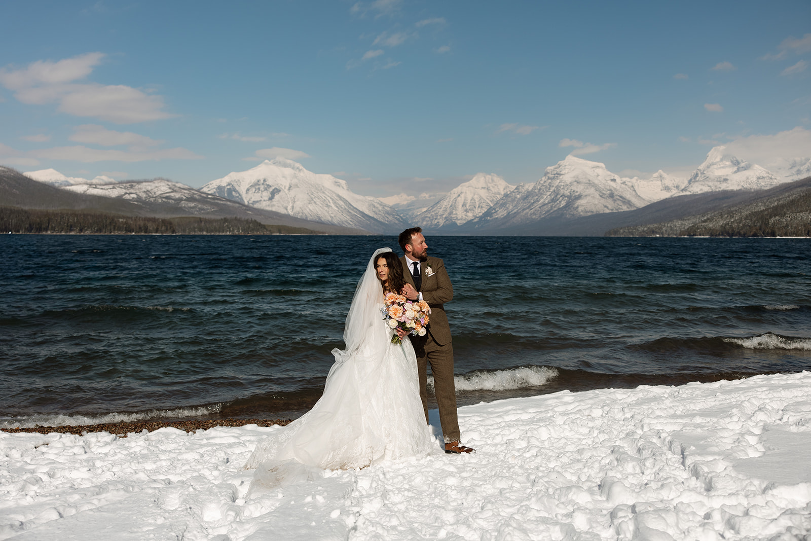 A winter elopement at Lake McDonald with a couple standing by a snowy lakeshore surrounded by mountains