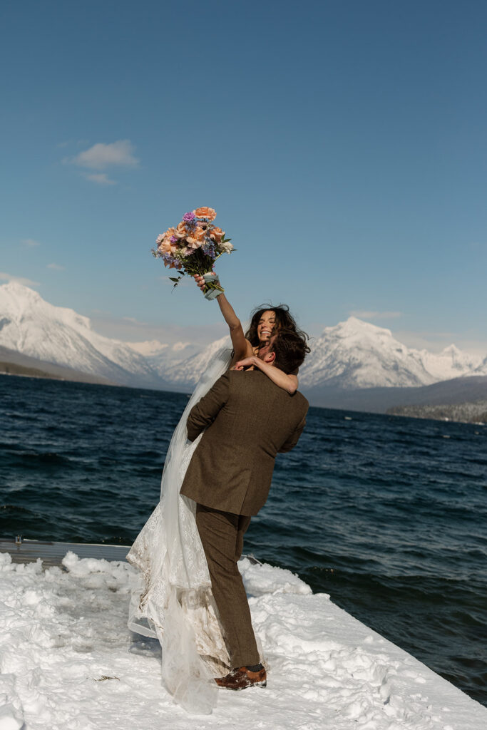 A winter elopement at Lake McDonald with a couple standing by a snowy lakeshore surrounded by mountains
