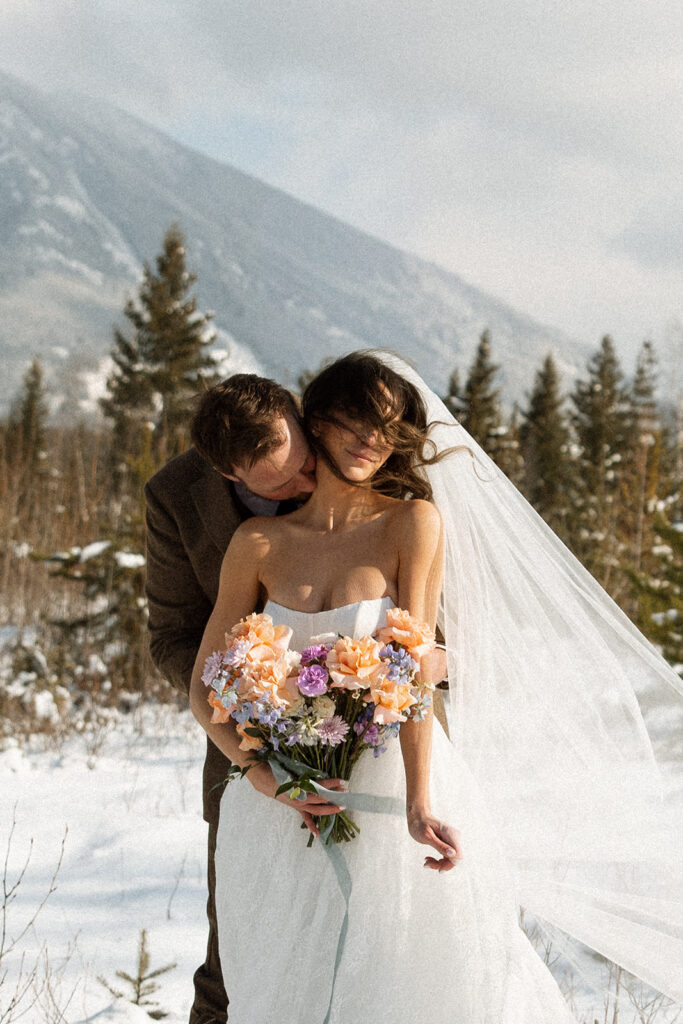 A winter elopement at Lake McDonald with a couple standing by a snowy lakeshore surrounded by mountains
